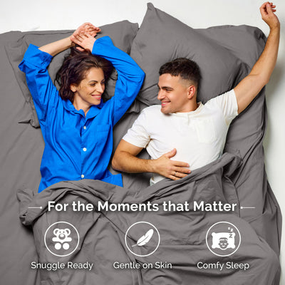 Couple lying on gray sheets, smiling and relaxing in bed, with pillows and bedding visible.