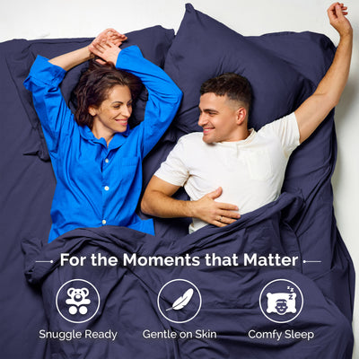 A woman in a blue shirt and a man in a white shirt relax on a bed with navy sheets and pillows.