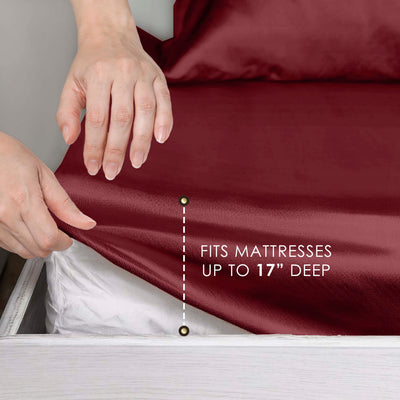 Hands adjusting a deep red satin sheet on a mattress, indicating it fits mattresses up to 17 inches deep.