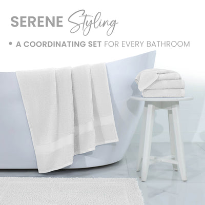 White bath towels hang over a bathtub, with a stack of folded towels on a white stool beside a textured bath rug.