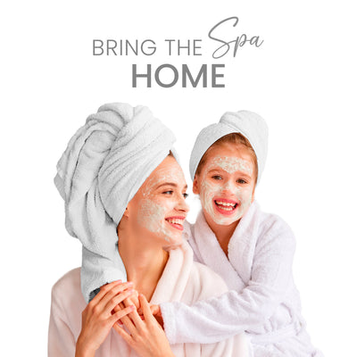 Two people with towels on their heads and facial masks, wearing white robes, smiling together.