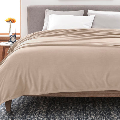 Soft beige fleece blanket draped over a bed with white and gray pillows, beside a nightstand with a clock and flower vase.