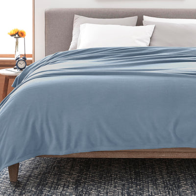 Soft blue fleece blanket draped over a bed with white pillows and a nightstand featuring a clock and a flower vase.