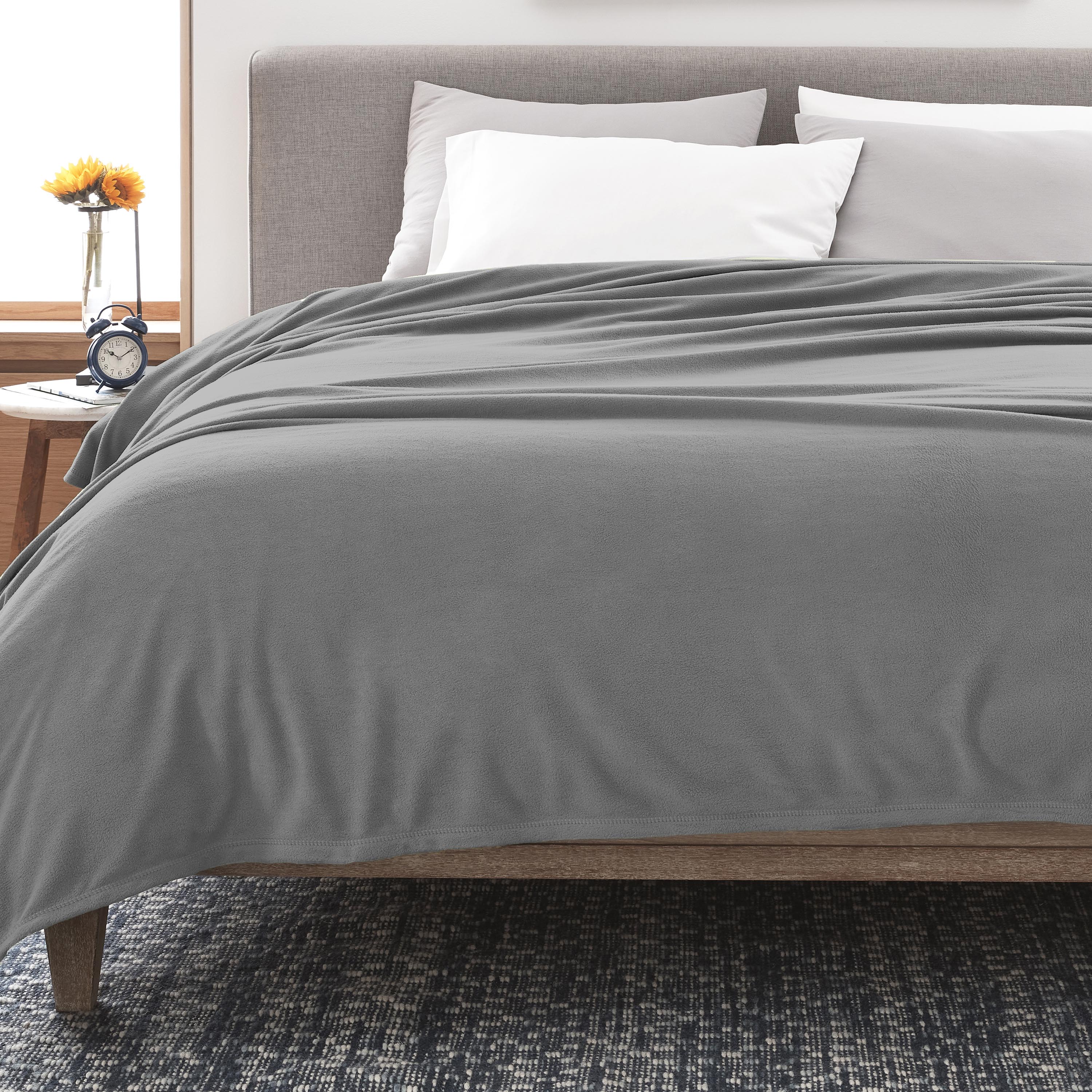 Gray fleece blanket draped over a bed with white pillows and a wooden nightstand featuring a clock and a vase with flowers.
