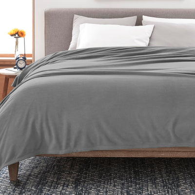 Gray fleece blanket draped over a bed with white pillows and a wooden nightstand featuring a clock and a vase with flowers.