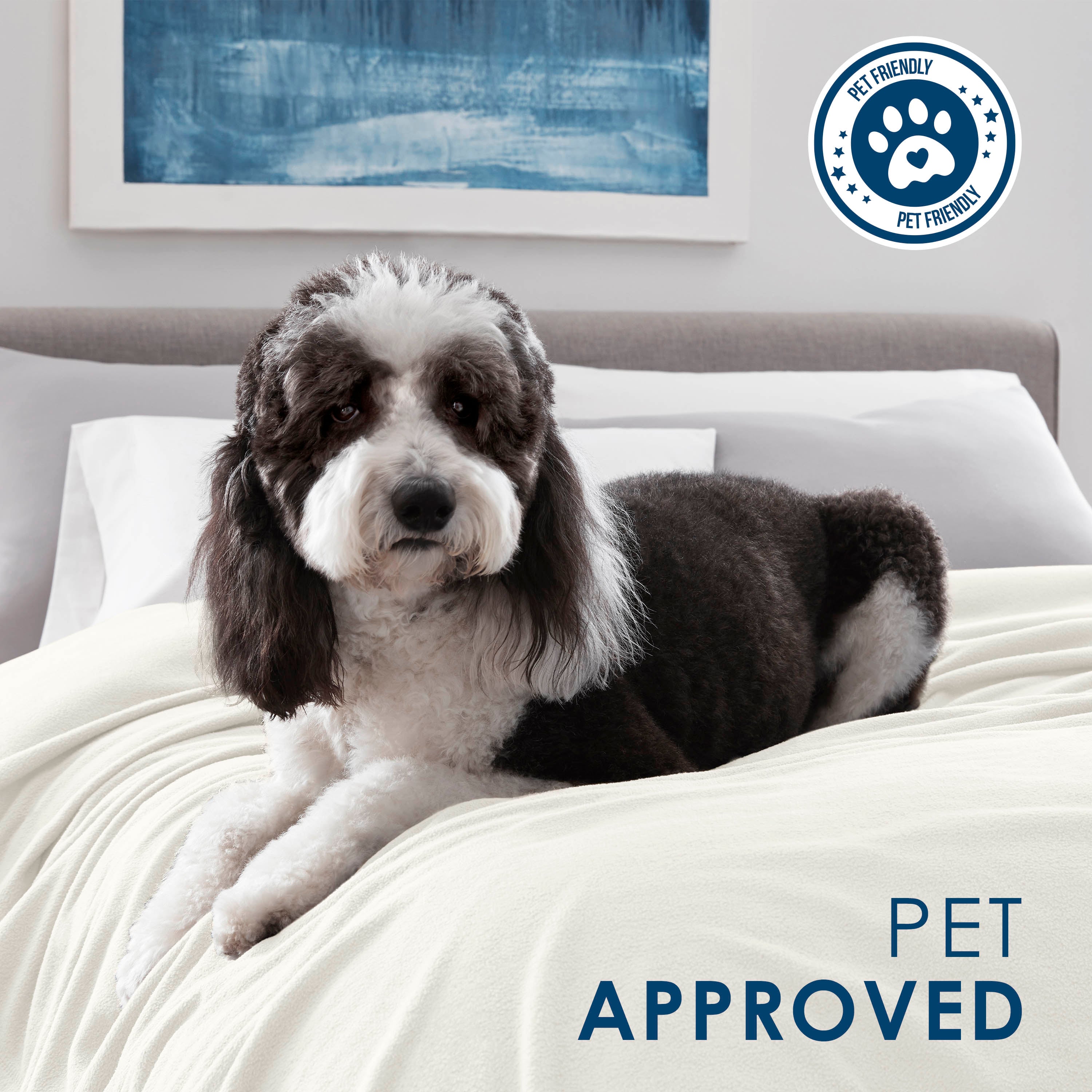 A dog rests on a cream-colored fleece blanket on a bed with white pillows and a blue abstract painting in the background.