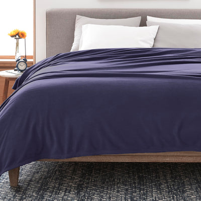 Navy fleece blanket draped over a bed with white pillows and a wooden nightstand featuring a clock and a flower vase.