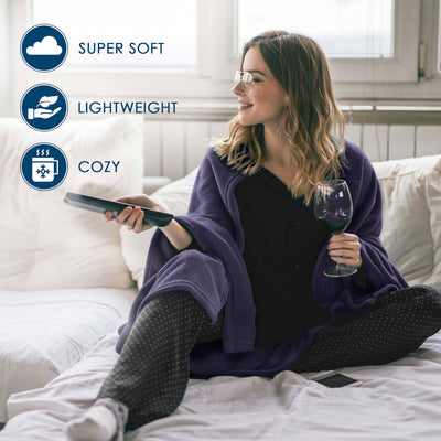 A woman sits on a bed wearing a purple fleece blanket, holding a remote and a glass of wine.