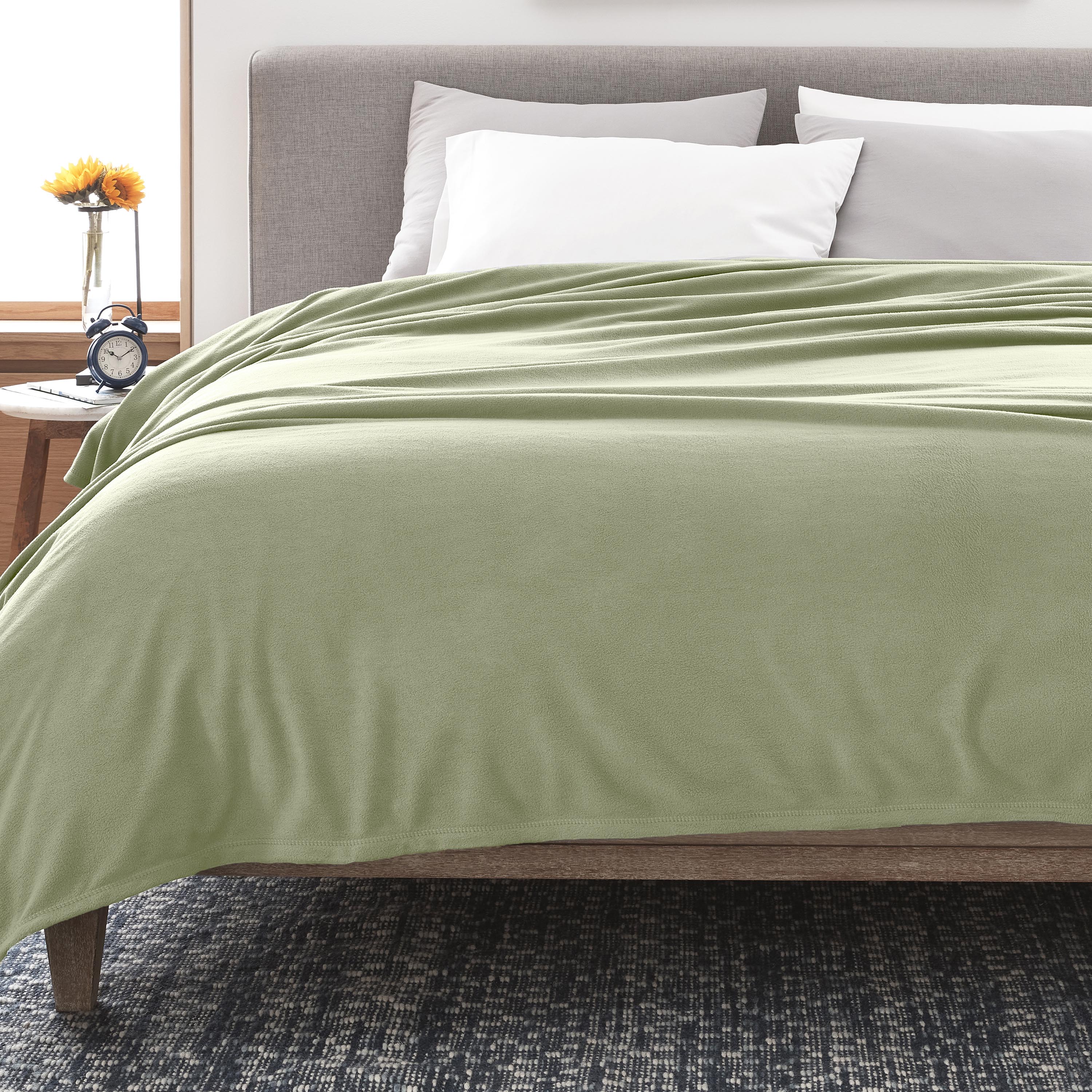 Light green fleece blanket draped over a bed with white pillows and a nightstand featuring a clock and a flower vase.