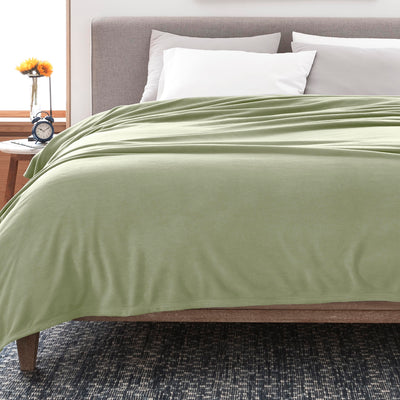 Light green fleece blanket draped over a bed with white pillows and a nightstand featuring a clock and a flower vase.