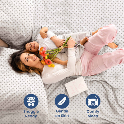 A woman and child lie on patterned sheets, with the woman holding flowers and smiling at the camera.
