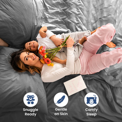 A woman and child lie on a gray bed, holding flowers and a card, with cozy pajamas and a relaxed atmosphere.