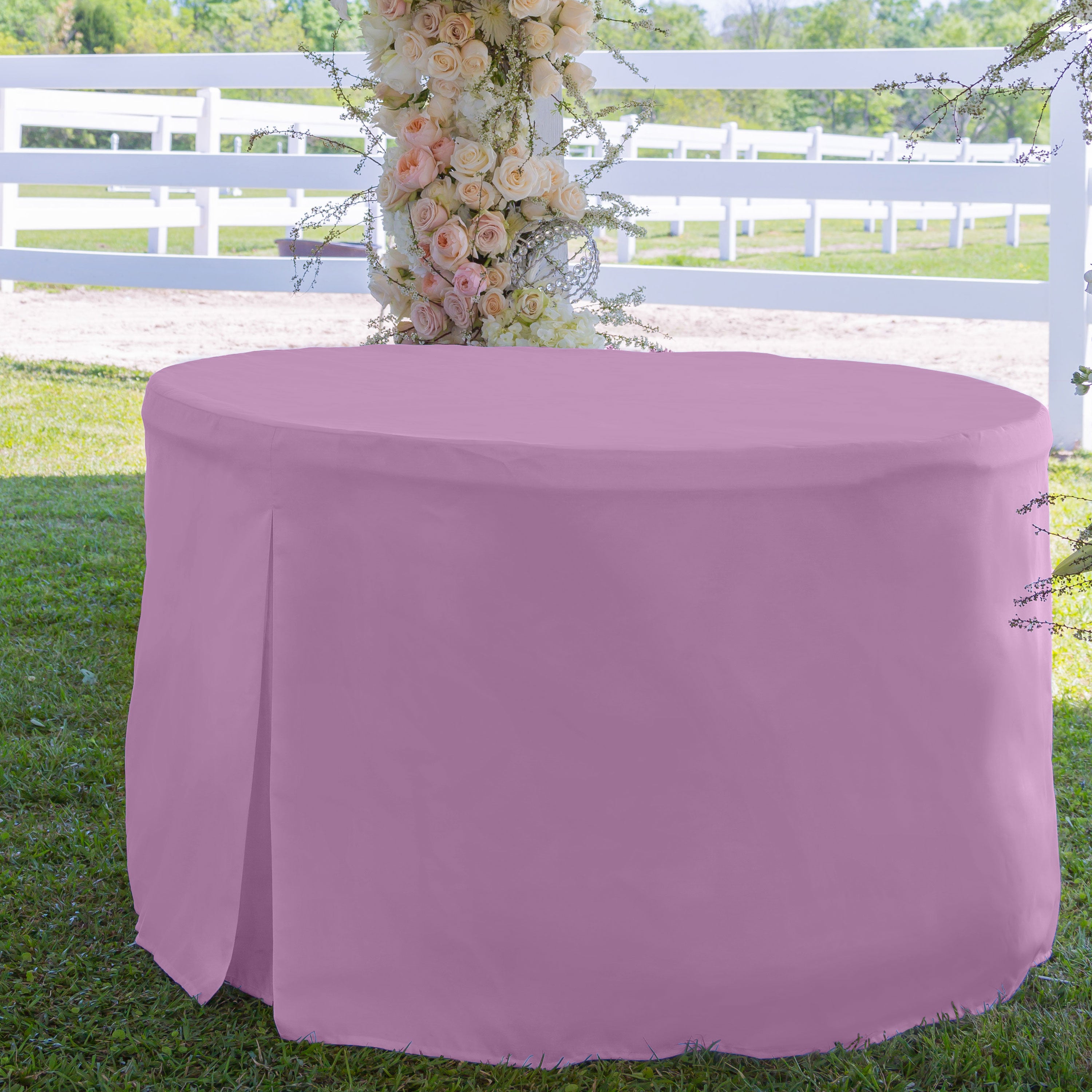 Round table covered with a solid pink tablecloth, set on grass with a floral backdrop.