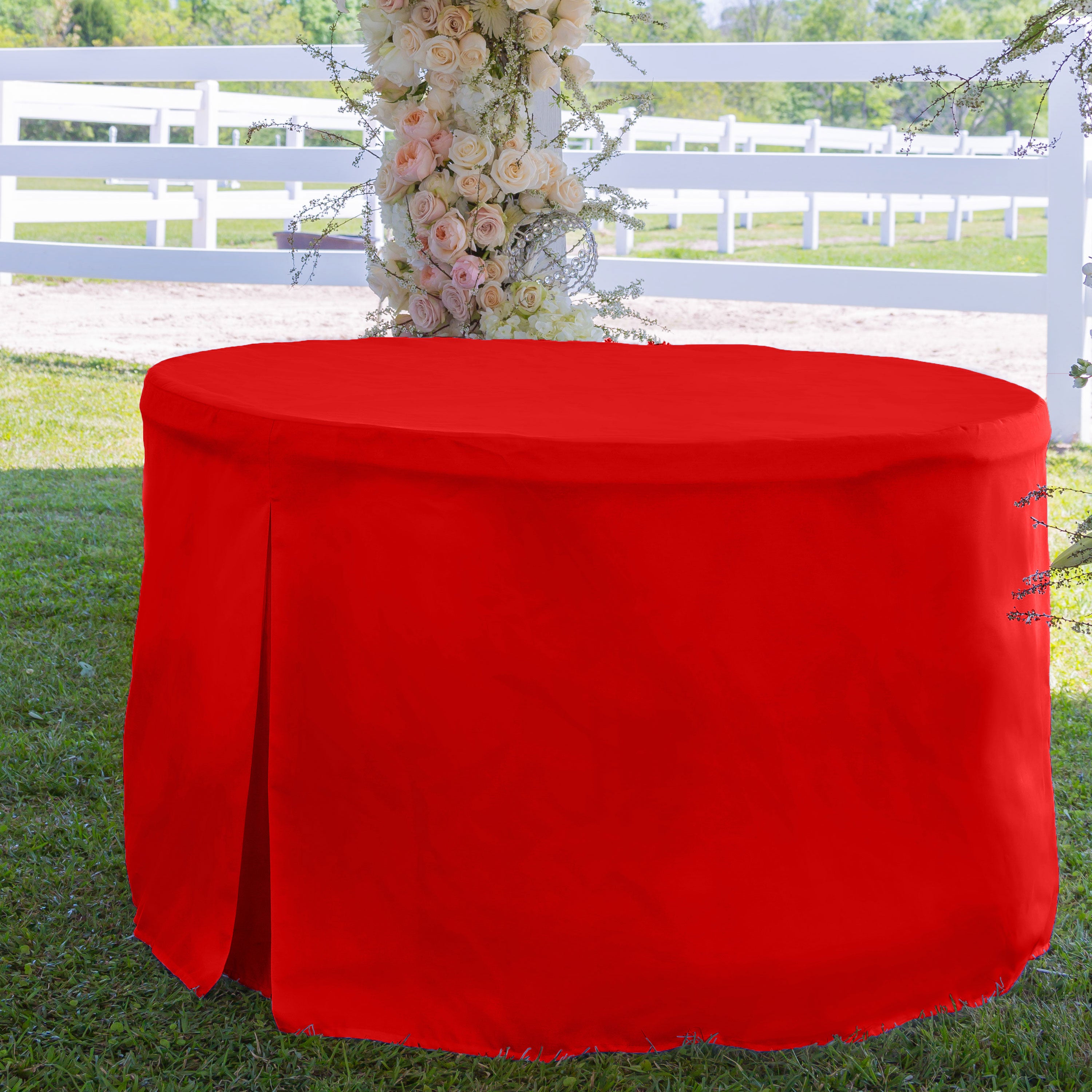 Red round table cover draped over a 60-inch table, set on grass with a floral backdrop.