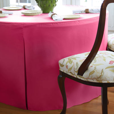 Round table covered with a pink tablecloth, set with plates and napkins, beside a floral-patterned chair.