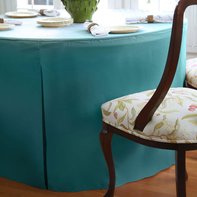 Teal round table cover drapes over a 60-inch table, with floral patterned chair visible beside it.