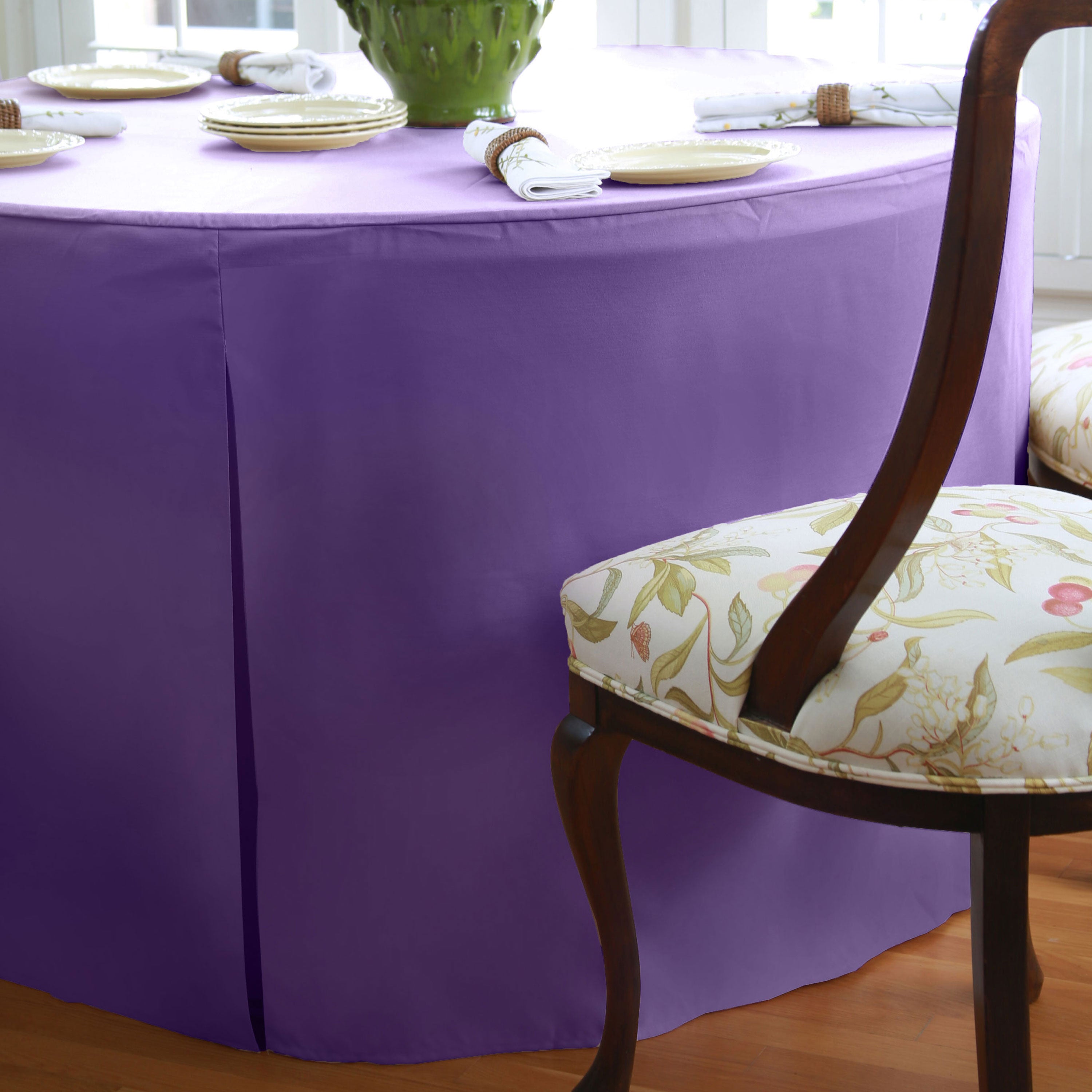 Round table covered with a purple tablecloth, with plates and napkins arranged on top, and a floral chair beside it.