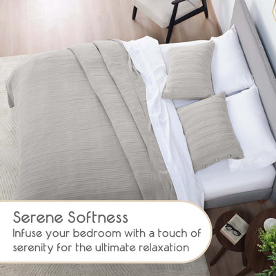 Light gray blanket and two matching pillows on a bed with white sheets and a light wood floor.
