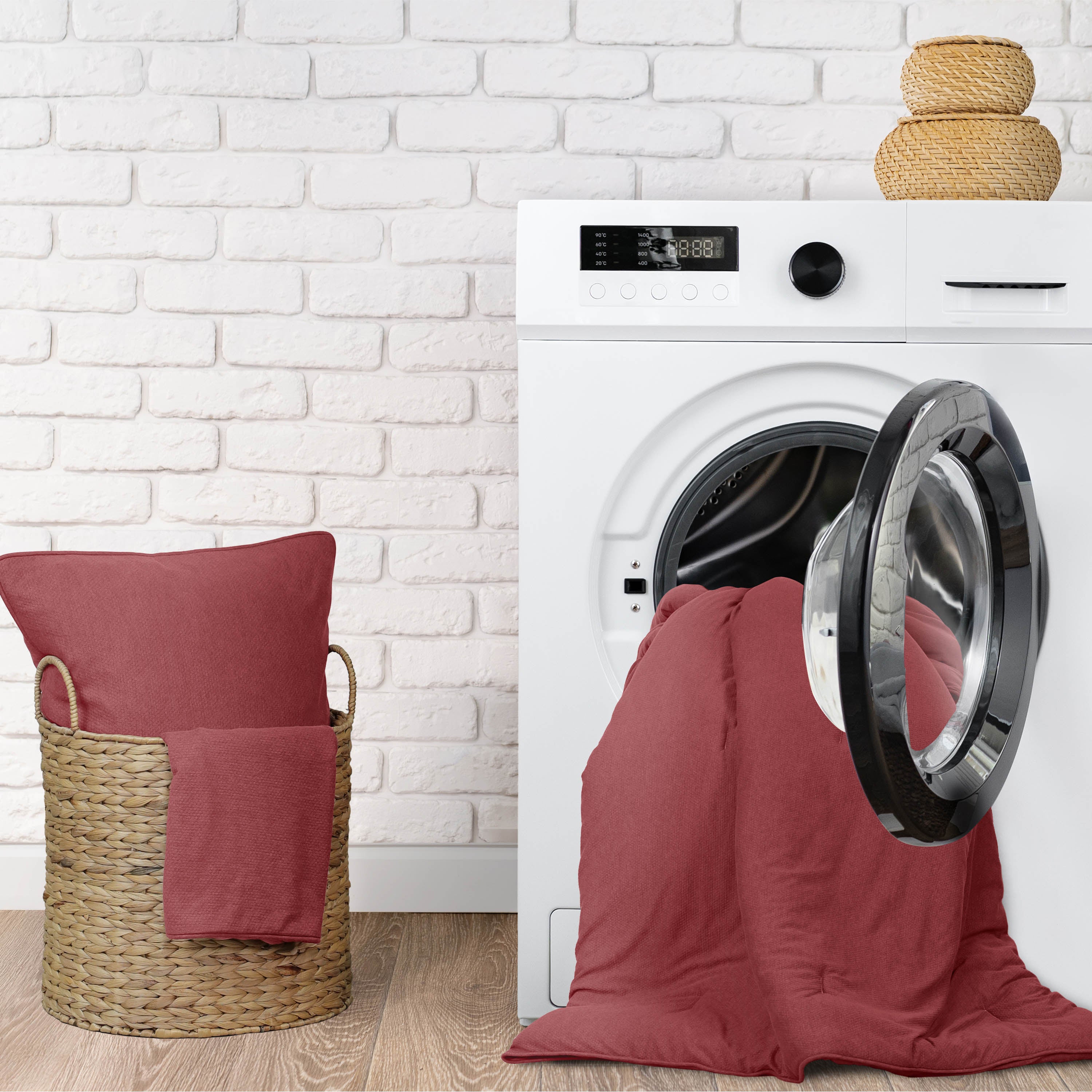 A washing machine with a comforter and pillow in red, alongside a woven basket containing a matching pillow.