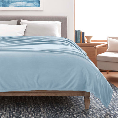 Light blue micro fleece blanket draped over a bed with gray headboard and white pillows, next to a wooden side table.