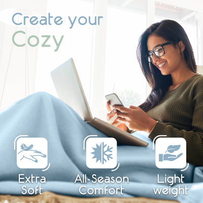 A woman sits on a couch using a laptop and phone, covered with a light blue micro fleece blanket.