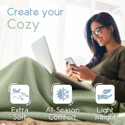 A woman sits on a couch, using a laptop and phone, covered with a light green micro fleece blanket.