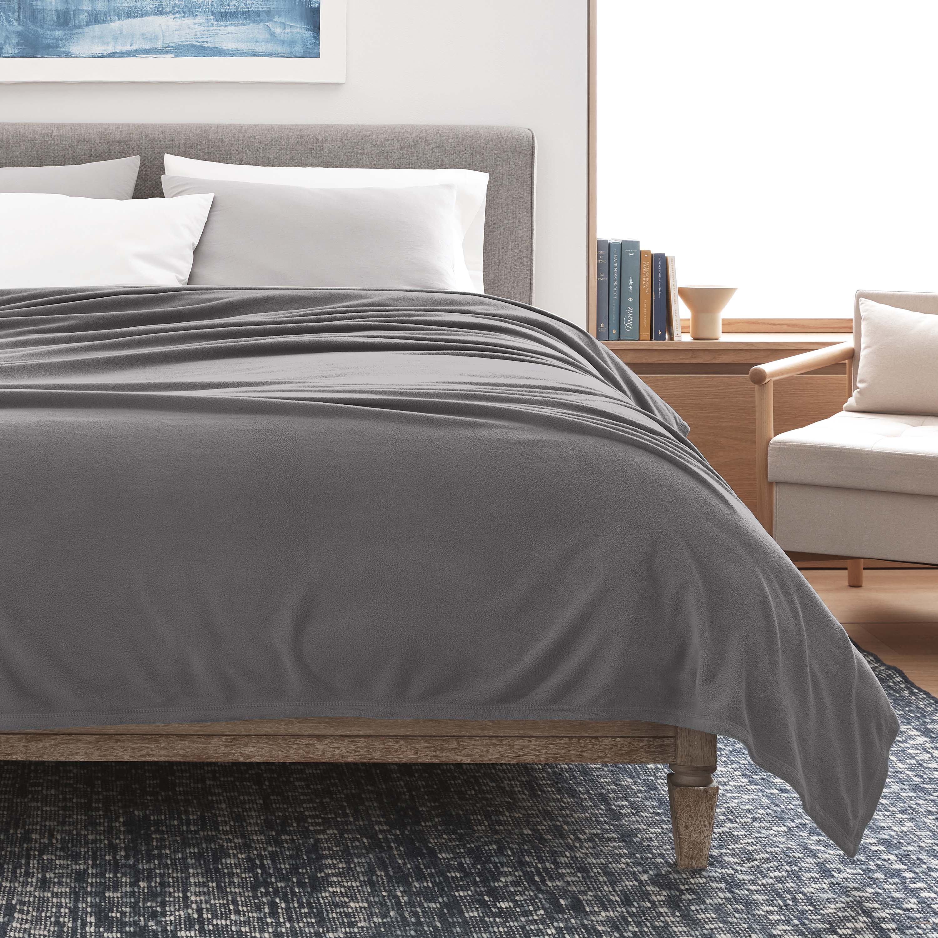 Gray micro fleece blanket draped over a bed with white pillows, a wooden nightstand, and a chair in the background.
