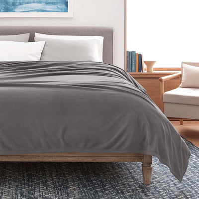 Gray micro fleece blanket draped over a bed with white pillows, a wooden nightstand, and a chair in the background.