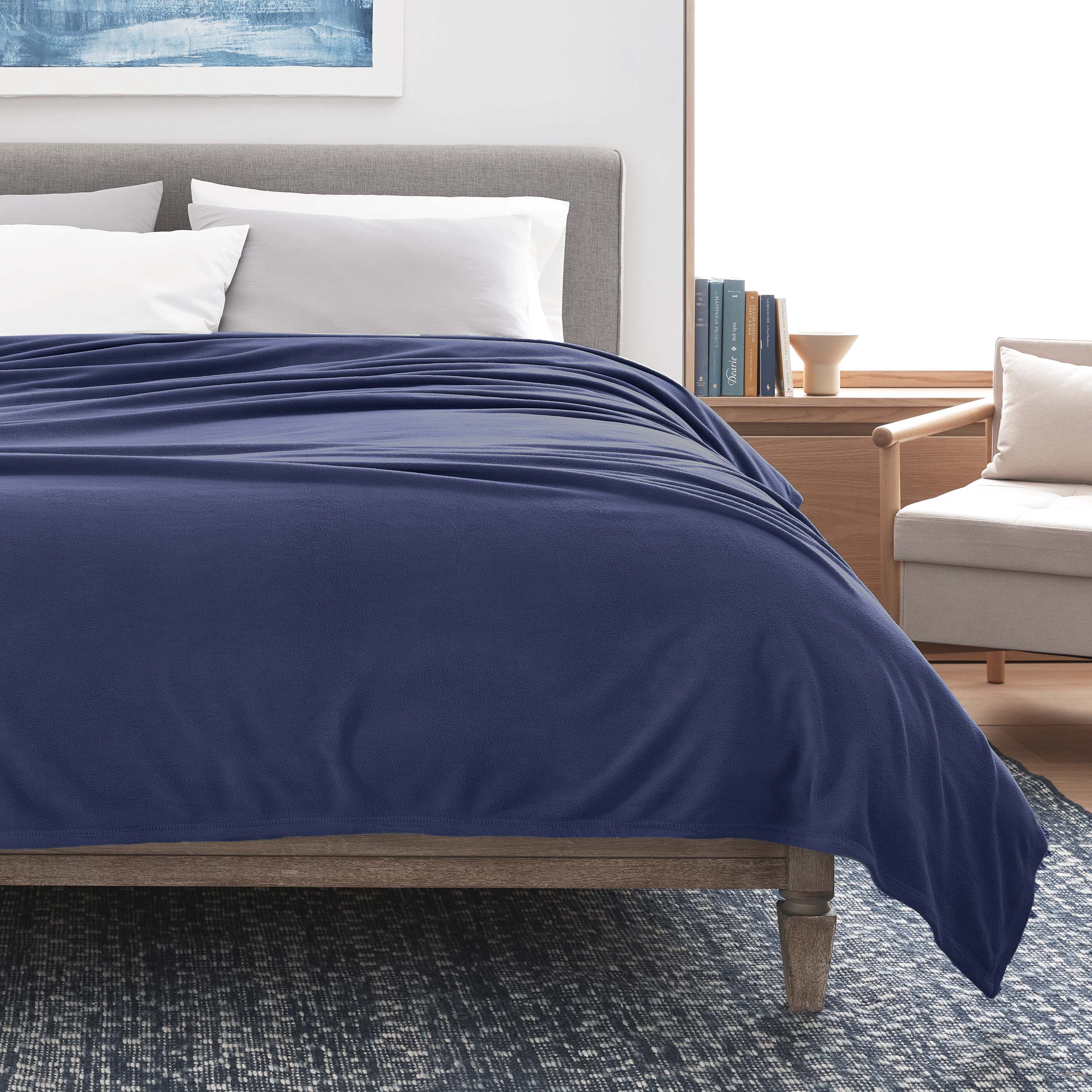 Navy micro fleece blanket draped over a bed with gray headboard and white pillows, beside a light-colored armchair.