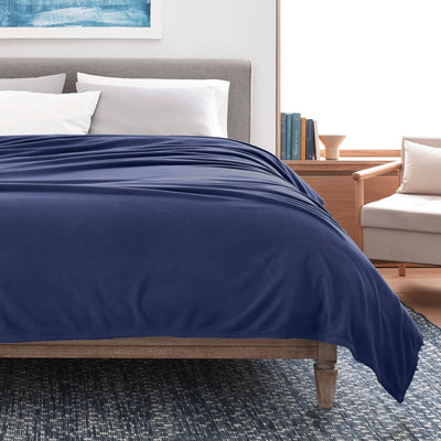 Navy micro fleece blanket draped over a bed with gray headboard and white pillows, beside a light-colored armchair.