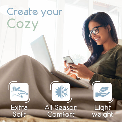 A woman sits on a couch with a laptop and smartphone, wrapped in a soft, light-colored blanket.