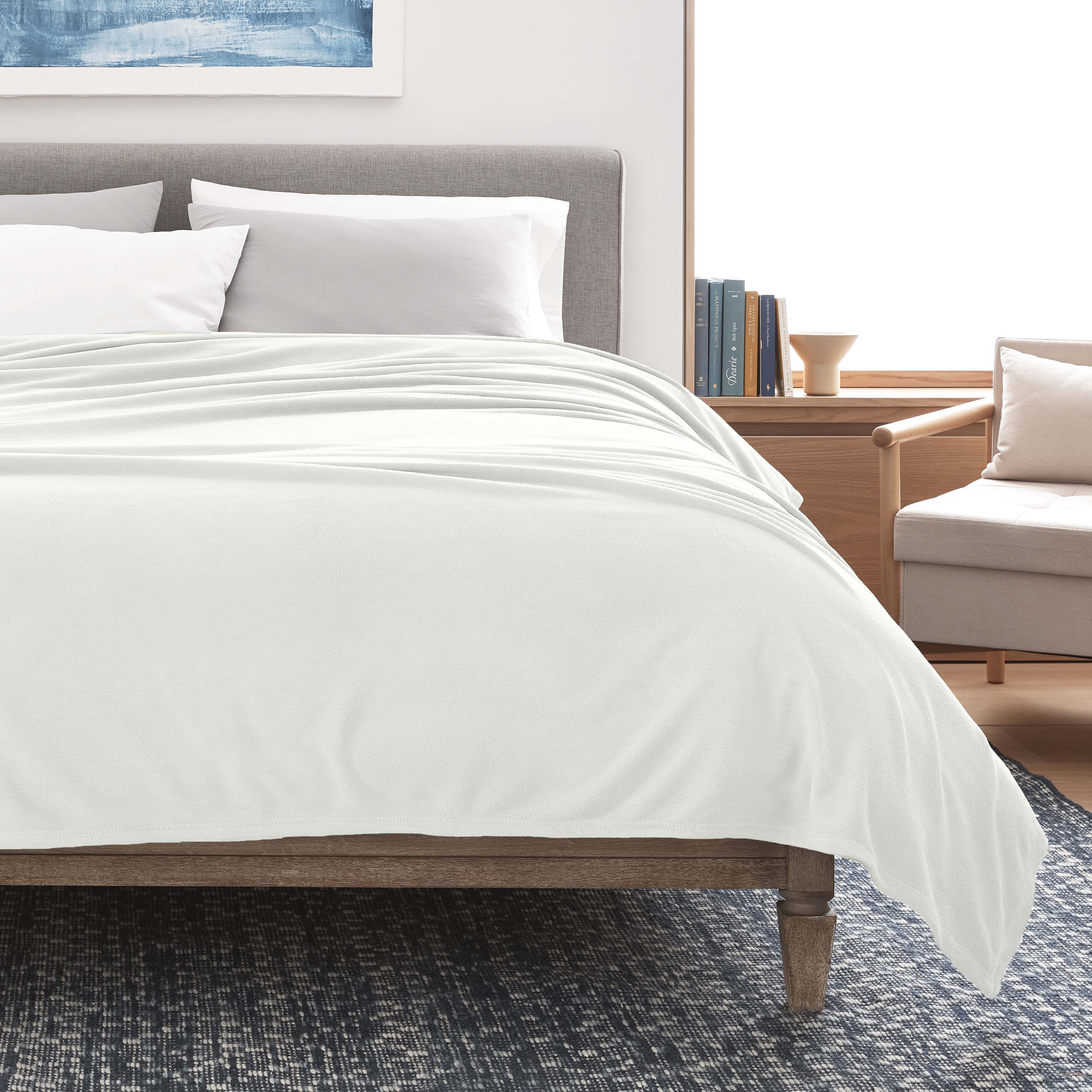 White micro fleece blanket draped over a bed with gray headboard and pillows, beside a light-colored armchair.