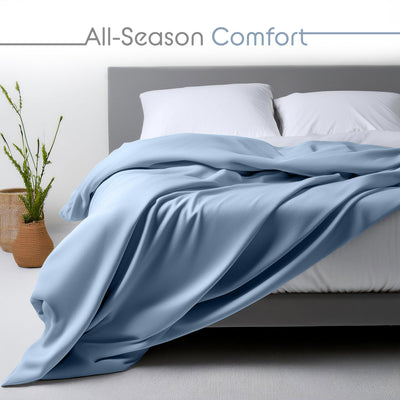 Light blue sheet set draped over a bed with white pillows and a plant in a woven basket nearby.