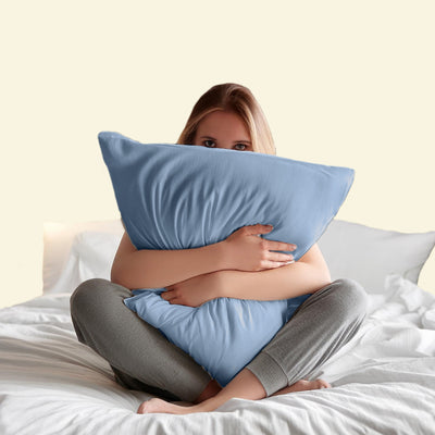 A person sits on a bed, hugging two blue pillows while wearing gray pants.