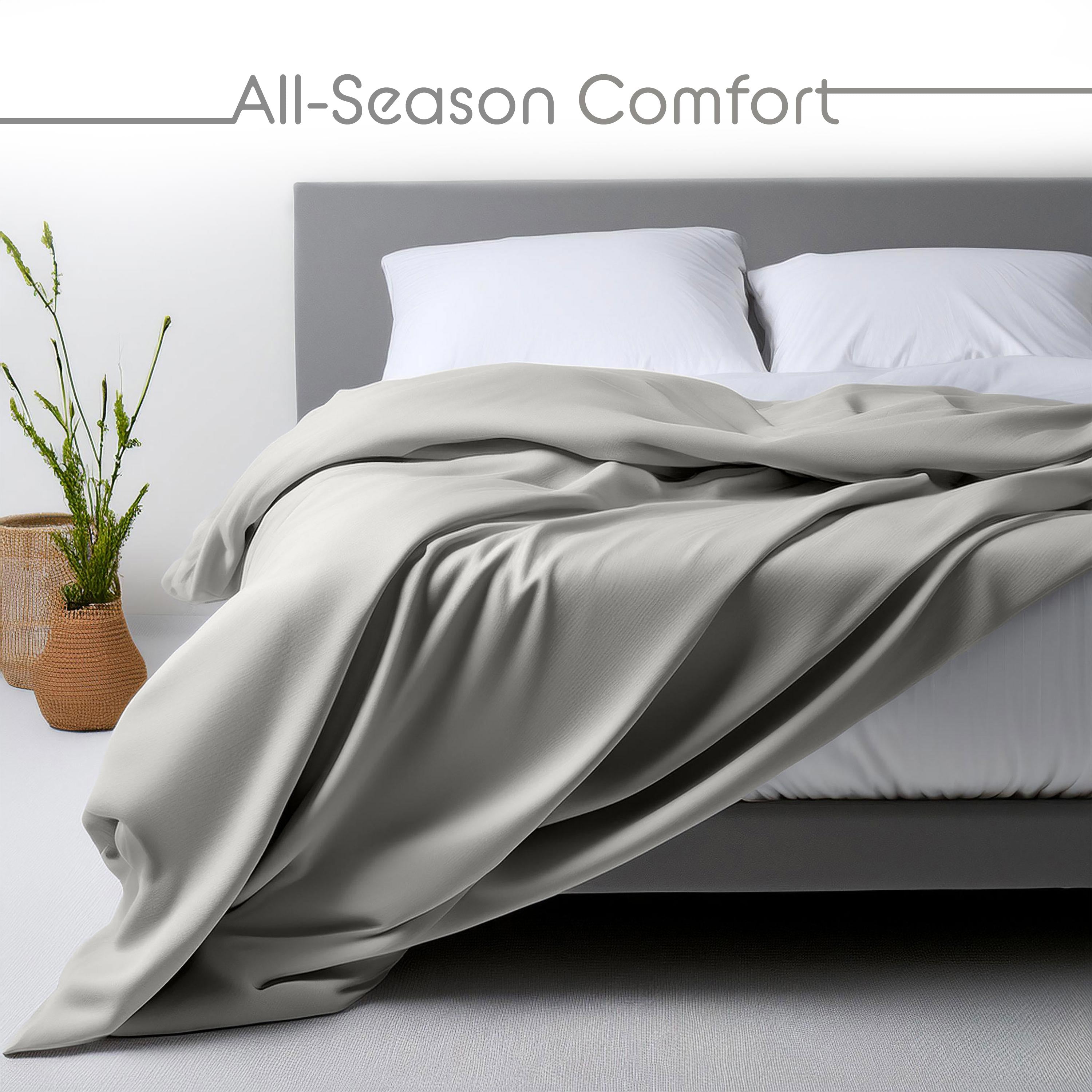 Light gray blanket draped over a bed with white pillows and a plant in a woven basket nearby.