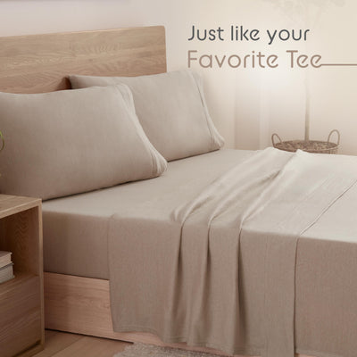 Beige bed with two large pillows and a soft sheet draped over the mattress, next to a wooden nightstand.