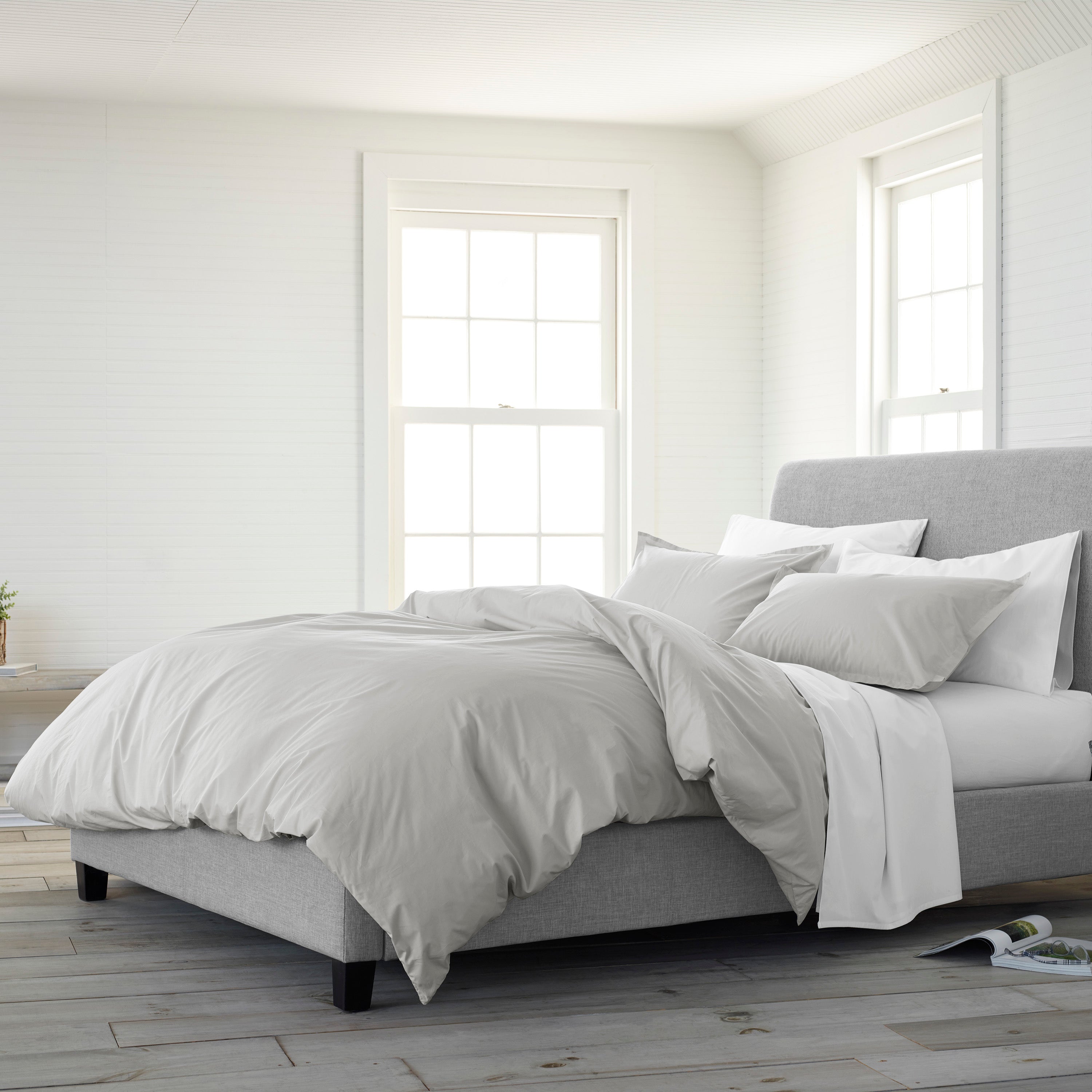 Solid gray comforter set on a bed with multiple pillows, in a bright room with large windows and wooden flooring.