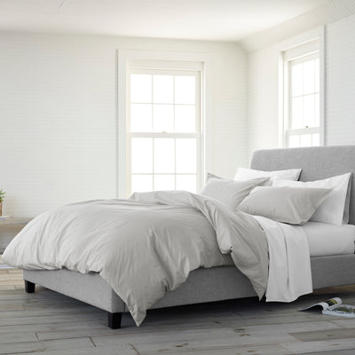 Solid gray comforter set on a bed with multiple pillows, in a bright room with large windows and wooden flooring.