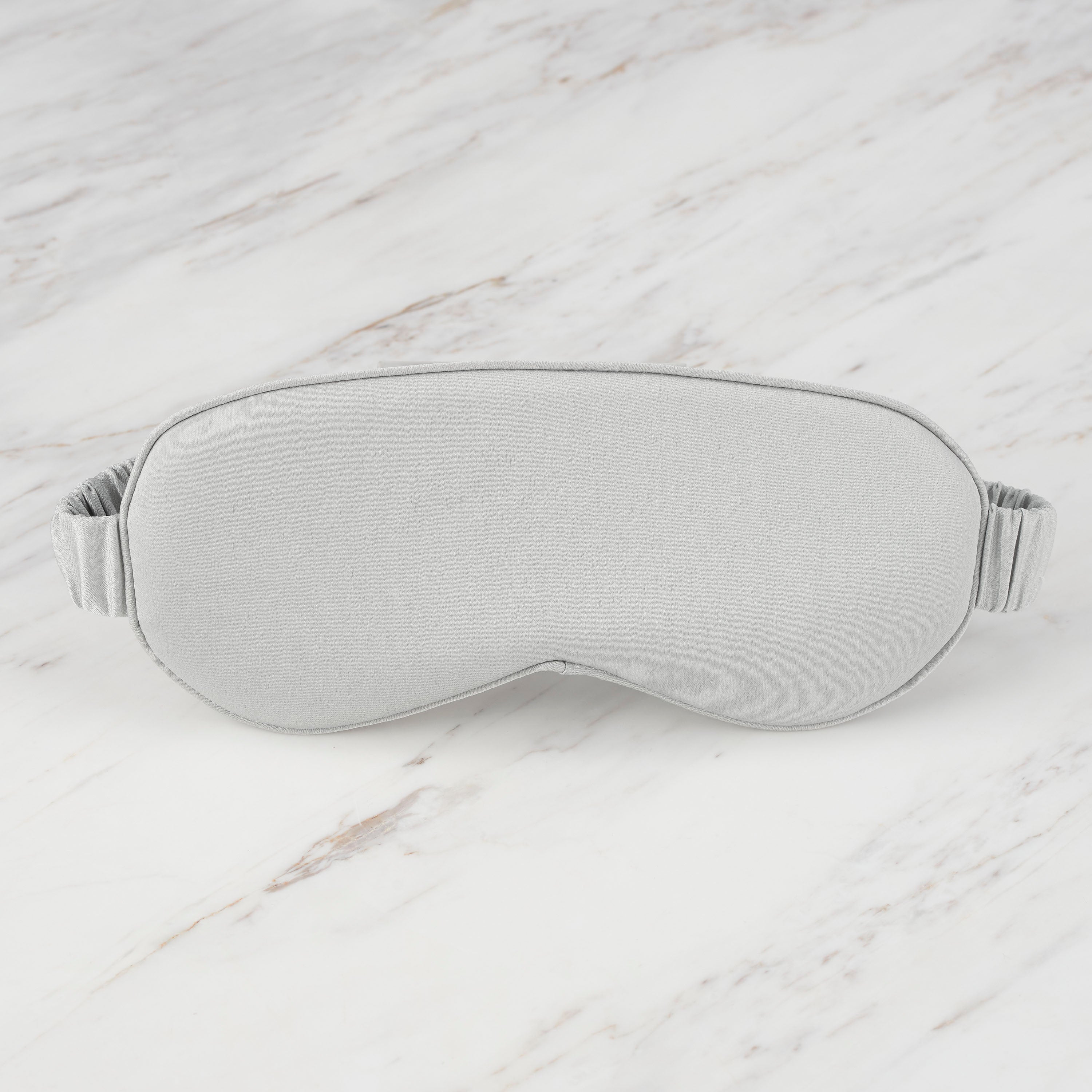 Gray satin eye mask with an elastic band, resting on a marble surface.
