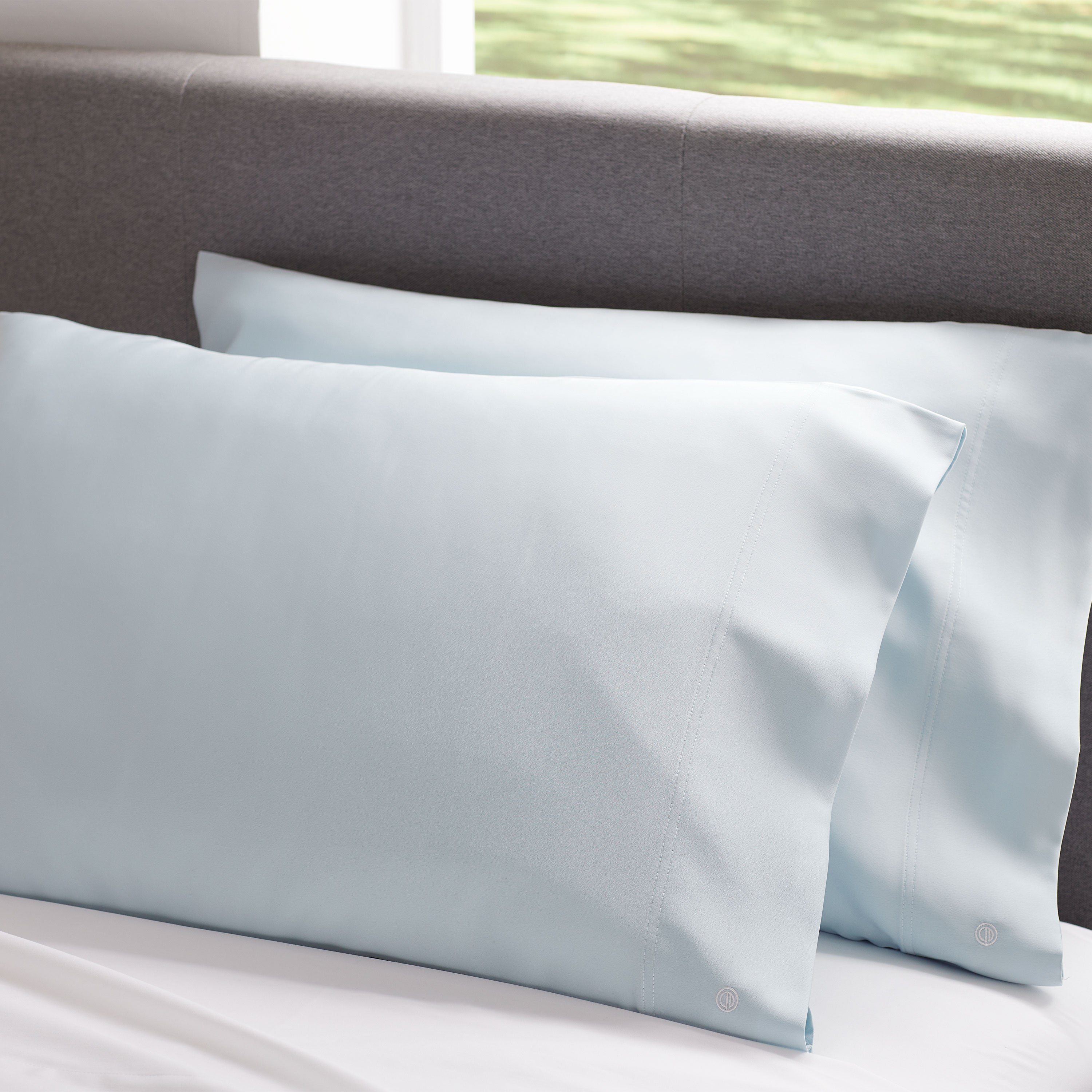 Light blue satin pillowcase pair resting on a white bed with a gray headboard in the background.