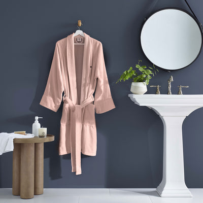 Light pink terry bath robe hanging on a hook next to a sink, with a plant and toiletries on a wooden table.