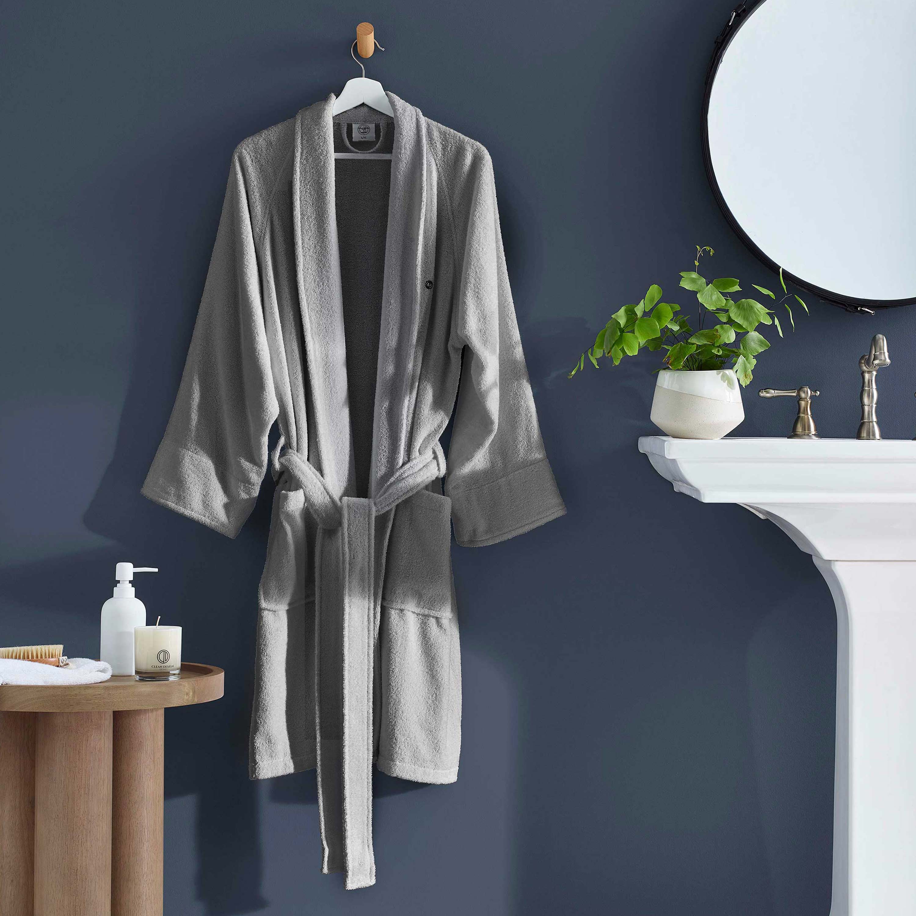 Gray terry bath robe with a belt, hanging on a hook against a dark wall, near a sink and potted plant.