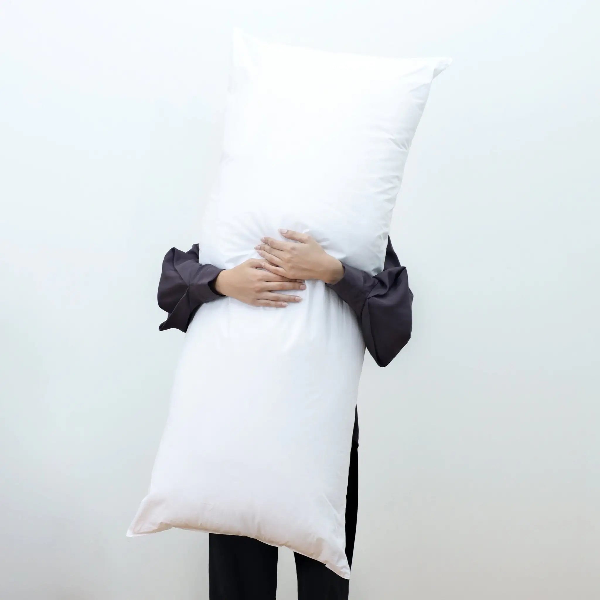 A person wearing a black long-sleeve shirt and black pants holds a large, white, rectangular body pillow against their face, obscuring it. The background is a plain, light off-white wall.