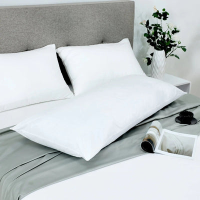 Three white pillows arranged on a bed with white and gray bedding, a gray upholstered headboard, a white side table, and a vase with greenery.