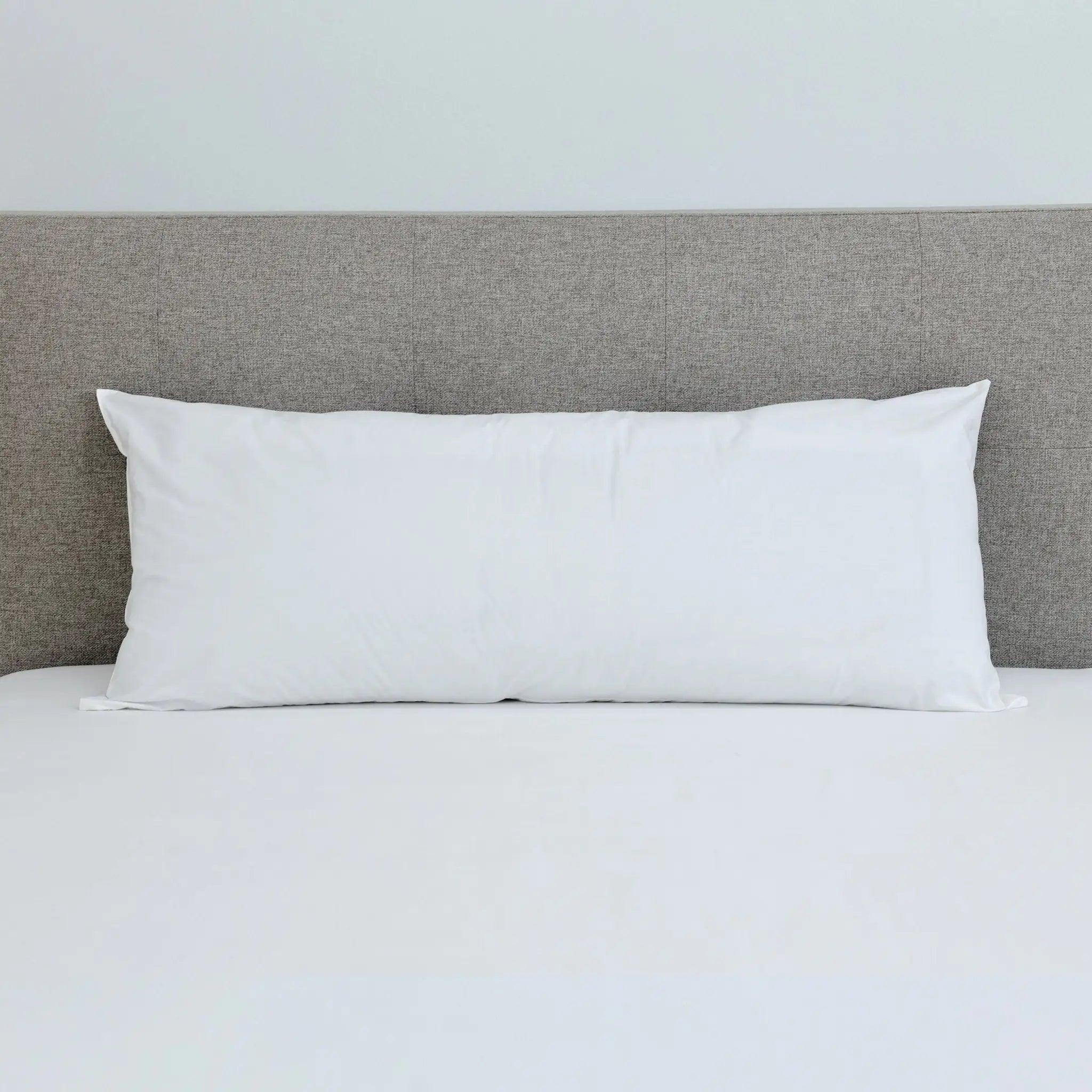 A white rectangular pillow resting on a bed with white sheets and a textured gray headboard against a light gray wall.