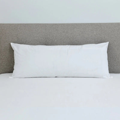 A white rectangular pillow resting on a bed with white sheets and a textured gray headboard against a light gray wall.