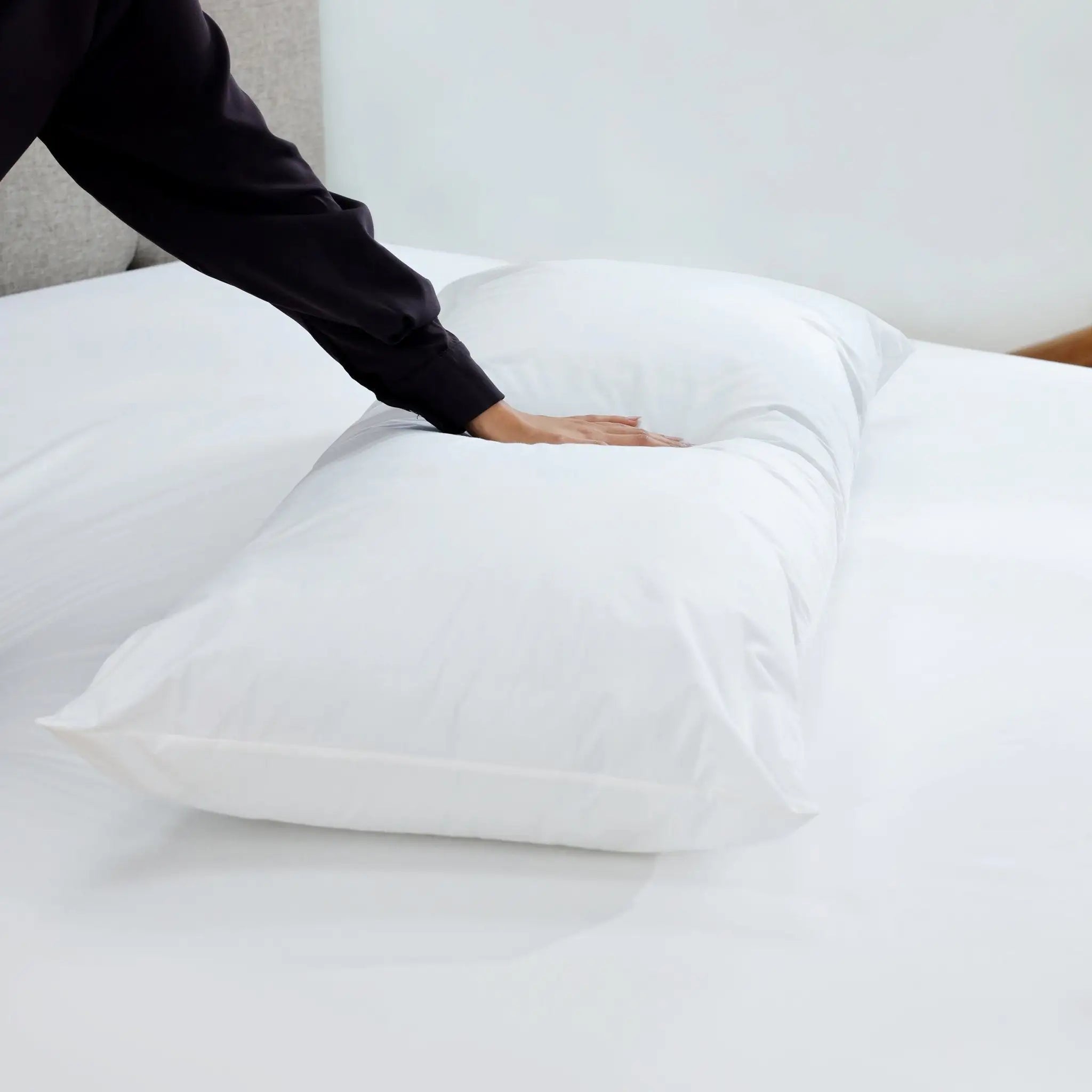 A white rectangular pillow resting on white bedding, with a person's hand pressing down on the front to demonstrate softness and comfort.