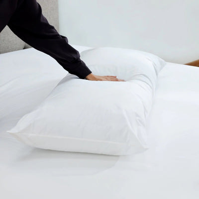 A white rectangular pillow resting on white bedding, with a person's hand pressing down on the front to demonstrate softness and comfort.