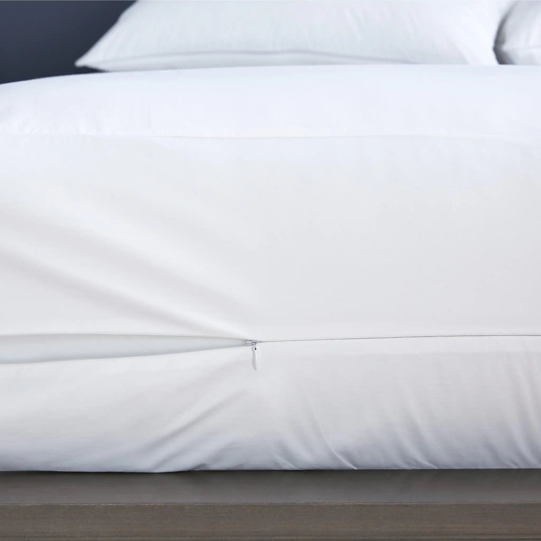 A close-up of a white mattress protector over the sides of a bed, with a small zipper seam visible near the bottom edge.