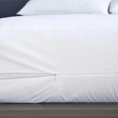 A close-up of a white mattress protector over the sides of a bed, with a small zipper seam visible near the bottom edge.